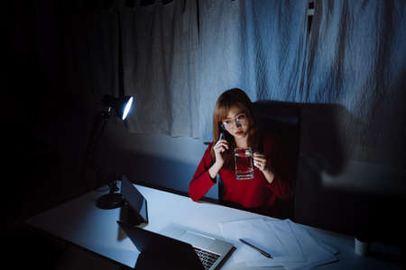 Asian woman working late time at night talking with smartphone, laptop computer home office unhappy emotion, Freelancer stay home business sleepless quarantine crisis coronavirus in dark room.の写真素材
