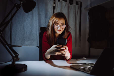 Asian woman working late time at night talking with smartphone, laptop computer home office happy relaxing, Freelancer stay home business quarantine sleepless crisis coronavirus stay home dark room.の写真素材