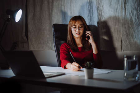 Asian woman working late time at night talking with smartphone, laptop computer home office happy relaxing, Freelancer stay home business quarantine sleepless crisis coronavirus stay home dark room.の写真素材