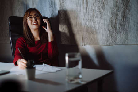 Asian woman working late time at night talking with smartphone, laptop computer home office happy relaxing, Freelancer stay home business quarantine sleepless crisis coronavirus stay home dark room.の写真素材