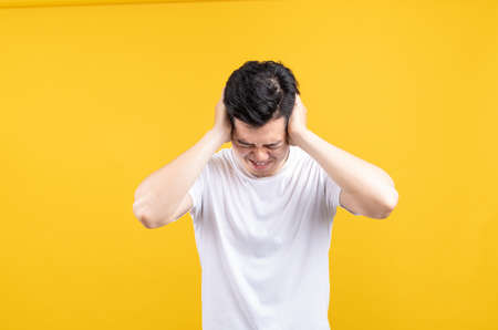 Attractive young asian man covering your ears loud noise shouting mouth angry and fury negative emotion wearing white t-shirt on yellow background isolated studio shot with copy spaceの写真素材