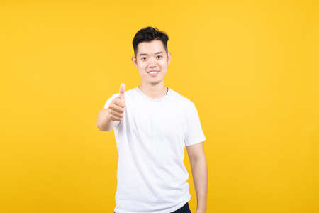 Attractive portrait happy young asian man smile laughing pointing at camera with copy space wearing white t-shirt on yellow background isolated studio shot.の写真素材