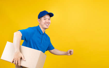 Delivery express of portrait asian male fast deliver to receiver carrying carton shipping buying online order wearing blue uniform on yellow background isolated studio shot.の写真素材