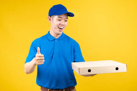 Delivery express of pizzaman asian male showing box delicious fast deliver to receiver shipping buying online order wearing blue uniform on yellow background isolated studio shot.の写真素材