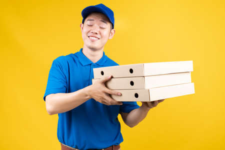 Delivery express of pizzaman asian male showing box delicious fast deliver to receiver shipping buying online order wearing blue uniform on yellow background isolated studio shot.の写真素材