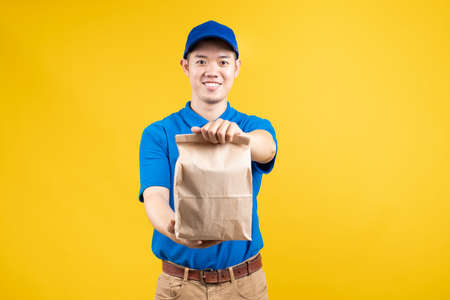 Delivery asian male showing food in paper bag delicious fast deliver to receiver shipping buying online order wearing blue uniform on yellow background isolated studio shot.の写真素材