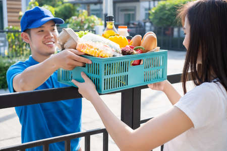 Food Deliver Asian man in blue uniform give fruit and vegetable to receiver customer front house, fast express grocery service when crisis coronavirus, covid19 new normal lifestyle concept.の写真素材