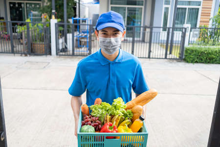 Food Deliver Asian man wearing mask in uniform give fruit and vegetable to receiver customer front house, fast express grocery service when crisis coronavirus, covid19 new normal lifestyle.の写真素材