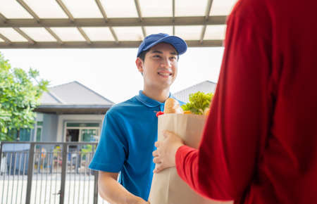 Food Deliver Asian man in blue uniform give fruit and vegetable to receiver customer front house, fast express grocery service when crisis coronavirus, covid19 new normal lifestyle concept.の写真素材