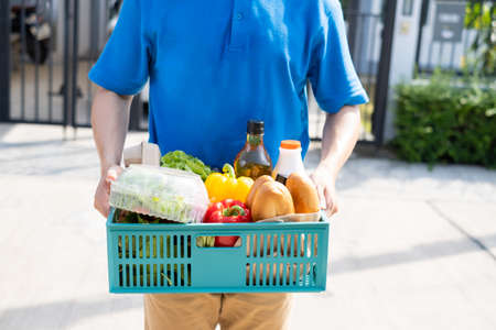 Food Deliver Asian man in blue uniform give fruit and vegetable to receiver customer front house, fast express grocery service when crisis coronavirus, new normal lifestyle concept.の写真素材
