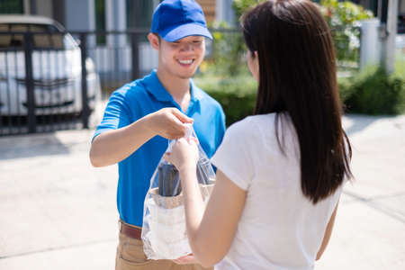 Food Delivery man wearing mask handing plastic bags shopping online from supermarket to female client service customer front house, express grocery when coronavirus covid19 new normal lifestyle.の写真素材