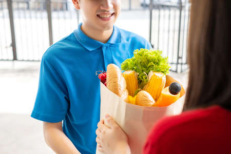 Food Delivery man in green uniform give fruit and vegetable shopping supermarket to receiver woman customer front house, express grocery service when crisis coronavirus, covid19 new normal lifestyle.の写真素材