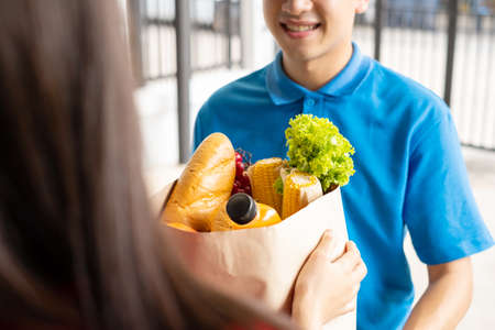 Food Delivery man in green uniform give fruit and vegetable shopping supermarket to receiver woman customer front house, express grocery service when crisis coronavirus, covid19 new normal lifestyle.の写真素材