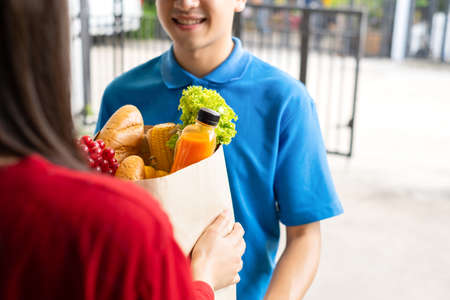 Food Delivery man in green uniform give fruit and vegetable shopping supermarket to receiver woman customer front house, express grocery service when crisis coronavirus, covid19 new normal lifestyle.の写真素材