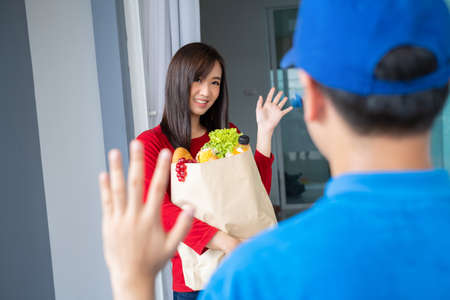 Food Deliver Asian man in blue uniform give fruit and vegetable to receiver customer front house, fast express grocery service when crisis coronavirus, covid19 new normal lifestyle concept.の写真素材