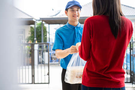 Food Delivery man wearing mask handing plastic bags shopping online from supermarket to female client service customer front house, express grocery when coronavirus covid19 new normal lifestyle.の写真素材