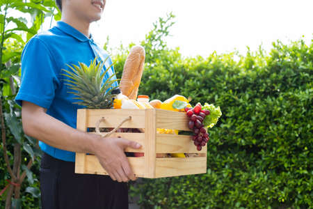 Food Deliver Asian man in blue uniform give fruit and vegetable to receiver customer front house, fast express grocery service when crisis coronavirus, new normal lifestyle concept.の写真素材