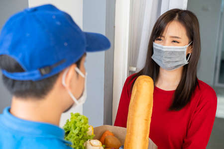 Food Delivery man wearing mask give fruit and vegetable shopping supermarket to receiver woman customer front house, express grocery service when crisis coronavirus, new normal lifestyle.の写真素材