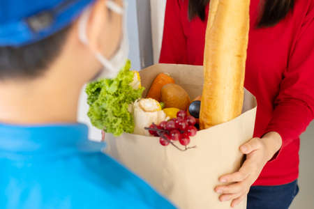 Food Delivery man wearing mask give fruit and vegetable shopping supermarket to receiver woman customer front house, express grocery service when crisis coronavirus, new normal lifestyle.の写真素材
