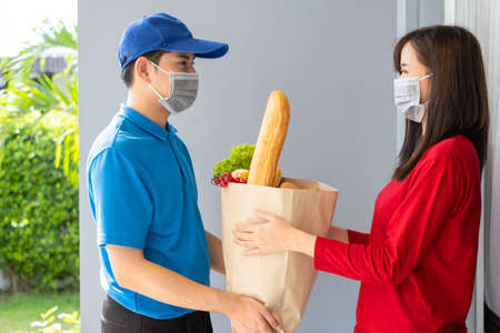 Food Delivery man wearing mask give fruit and vegetable shopping supermarket to receiver woman customer front house, express grocery service when crisis coronavirus, new normal lifestyle.の写真素材