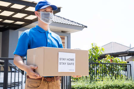 Delivery service male in uniform give cardboard box package to receiver customer client front home, fast express offsite when crisis coronavirusの写真素材