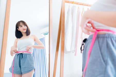 Beautiful young asian woman with a tape measure body perfect slim weight loss in front of a mirror people lifestyle at home in the morning.の写真素材