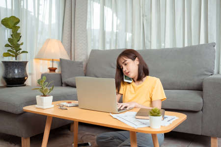 Young asian woman working on laptop computer and using cellphone while sitting at the living room, Freelance girl working at home, work online, home office.の写真素材