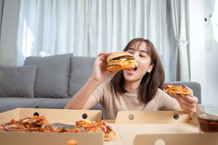 Close up asian woman takeaway eating junk food hamburger and pizza close-up shot in living room at home,  Fast food delivery service safe and stop coronavirus spread by social distancing concept.の写真素材