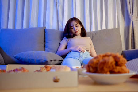 Young asian woman takeaway eating full junk food sitting on sofa and watch television in living room at home Fast food delivery service, She is already tried to sleep.の写真素材