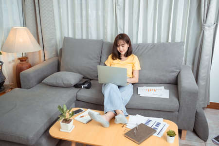 Young asian woman wearing headphones having video chat with friends using laptop camera while siting on couch at the living room, Online meeting in video call for social distancing.の写真素材