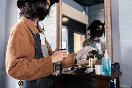 Male Barber cleaning equipment hair clippers with alcohol spray before hair cut he wearing face mask prevention business reopening after covonarirus lockdown, Men's hairstyling new normal lifestyle.の写真素材