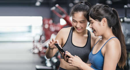Two sporty young asian women with friends relaxing after their workout at the gym talking and smiling in fitness, Warming up checking mobile smartphone watching healthy lifestyle with copy space.の写真素材