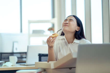 Business woman sitting on the chair eating pizza for lunch while working. She happy diverse tasting Italian food, takeaway pizza box meal at workplace with colleagues in office. Service concept.の写真素材