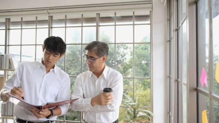Young asian businessmen holding paper documents to discuss information with mature businessman holding a cup of coffee. Creative business people in modern office. Two men discussing marketing plan.の写真素材