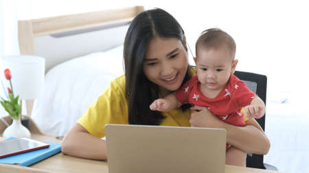Beautiful business asian mom is using a laptop and smiling while spending time with her cute baby at home. Young mother on maternity leave trying to freelance by the desk with toddler child.の写真素材