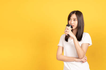 Happy asian young woman wearing white t-shirt standing and holding a coffee happy, smiling and cheerful at studio shot on yellow background. isolated famale portrait.の写真素材