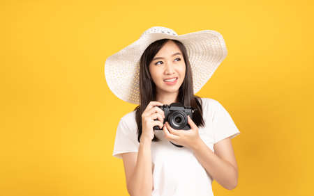 Happy smiling asian young girl photographer wearing hat and hold camera DSLR and photo camera ready to shoot at studio shot on yellow background with copy space.の写真素材