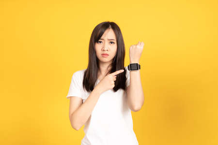 Asian young woman wearing white t-shirt standing. She looking on smart watch on hand put hand on cheek at studio shot on yellow background. People lifestyle concept.の写真素材