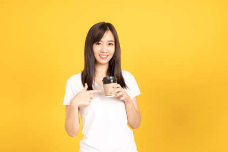 Happy asian young woman wearing white t-shirt standing and holding a coffee happy, smiling and cheerful at studio shot on yellow background. isolated famale portrait.の写真素材