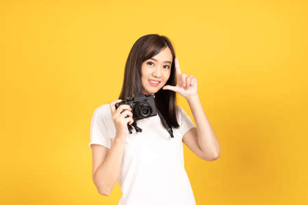 Happy smiling asian young girl photographer takes images with dslr camera on retro digital mirrorless photo camera ready to shoot at studio shot on yellow background.の写真素材