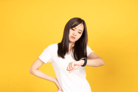 Asian young woman wearing white t-shirt standing. She looking on smart watch on hand put hand on cheek at studio shot on yellow background. People lifestyle concept.の写真素材
