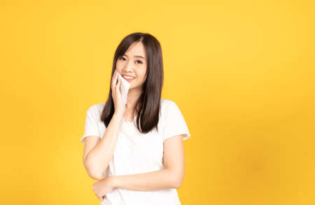 Asian young woman wearing white t-shirt standing and holding a tissue paper on her face female with hygiene paper at studio shot on yellow background. isolated famale portrait.の写真素材