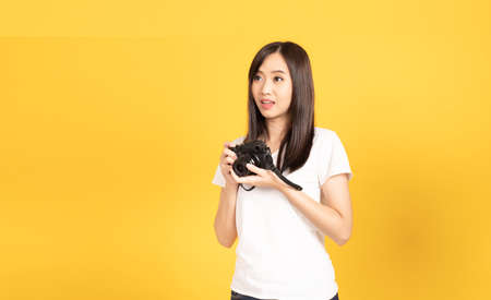 Happy smiling asian young girl photographer takes images with dslr camera on retro digital mirrorless photo camera ready to shoot at studio shot on yellow background.の写真素材
