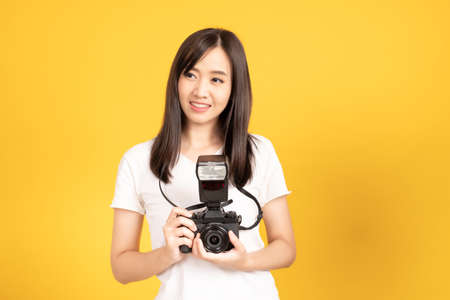 Happy smiling asian young girl photographer and looking viewfinder on retro digital mirrorless photo camera ready to shoot at studio shot on yellow background.の写真素材