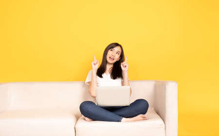 Young asian woman point finger on comfortable couch, using laptop at home, She is happy to spend some free time online while chatting with friends in social network, shopping or working online.の写真素材