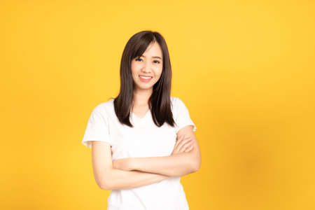 Happy asian young woman wearing white t-shirt standing with crossed arms ready to shoot at studio) shot on yellow background. isolated famale portrait with copy space.の写真素材