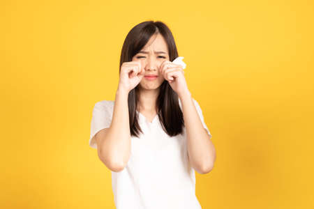 Asian young woman wearing white t-shirt standing and holding a tissue paper using her face wiping tears at studio shot on yellow background. isolated famale portrait.の写真素材