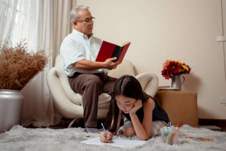 Asian handsome grandpa is holding a book while his granddaughter is drawing on a paper are smiling while spending time together sit on sofa in the living room at home. Enjoy family concept.の写真素材