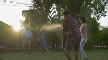 Happy asian senior grandpa pouring water from a hose. Funny family with his father playing with family having fun with spray of water in garden house in sunny backyard. Enjoy family holiday concept.の写真素材