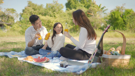 Happy family asian and little girl are drinking orange juice and have enjoyed ourselves together during picnicking on a picnic cloth in the green garden. Family enjoying sunny fall day in nature.の写真素材